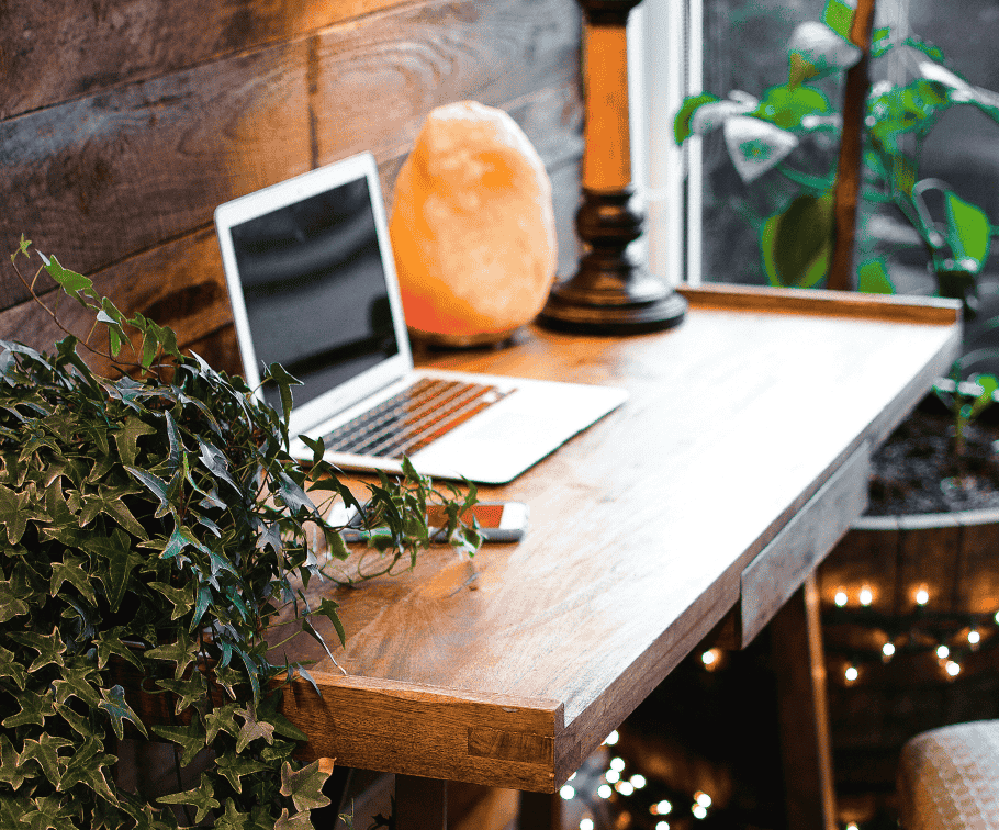 A small wooden work desk for a freelance transcriptionist with an open laptop and a Himalayan salt crystal lamp and a green hanging plant and a string of warm lights